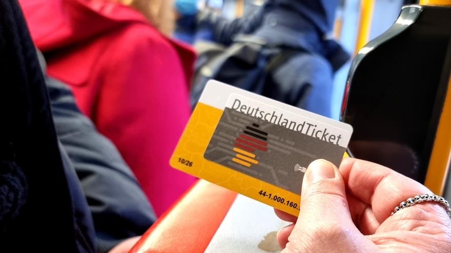 Eine Hand hält ein Deutschlandticket neben einen Stopp-Knopf an der Tür einer Straßenbahn